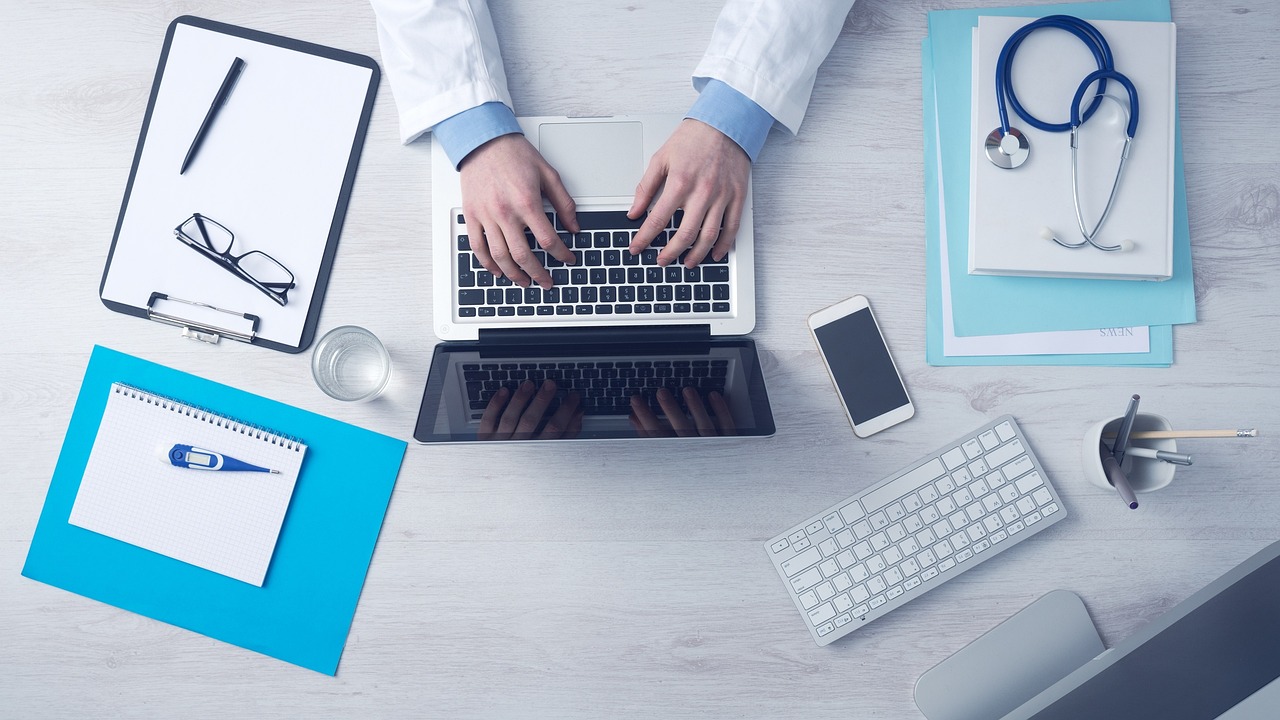 A doctor works at a tidy desk with a laptop, clipboard, and stethoscope