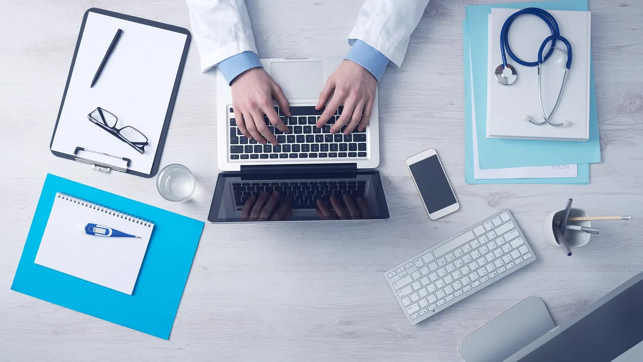 A desk with medical paperwork, a clipboard, and a laptop